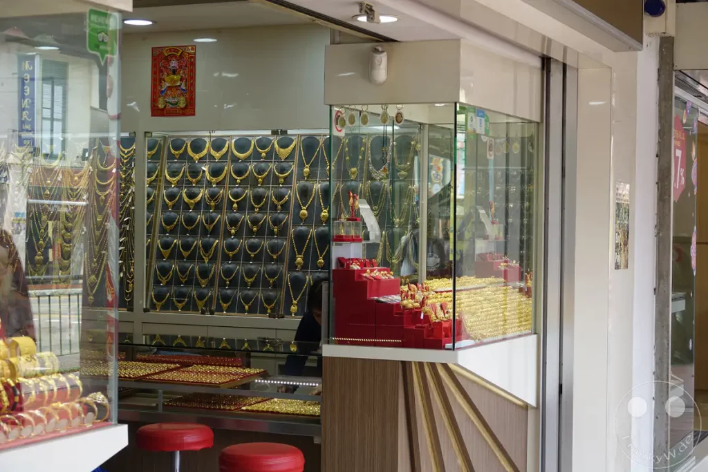 Eine Vielzahl von goldenen Halsketten und Armbändern wird in einem Juweliergeschäft in Singapur ausgestellt. Die glänzenden Schmuckstücke sind in Schaufenstern und auf roten Ständern arrangiert, um Kunden anzulocken. Die beleuchtete Auslage erzeugt eine Atmosphäre des Luxus und der Opulenz.