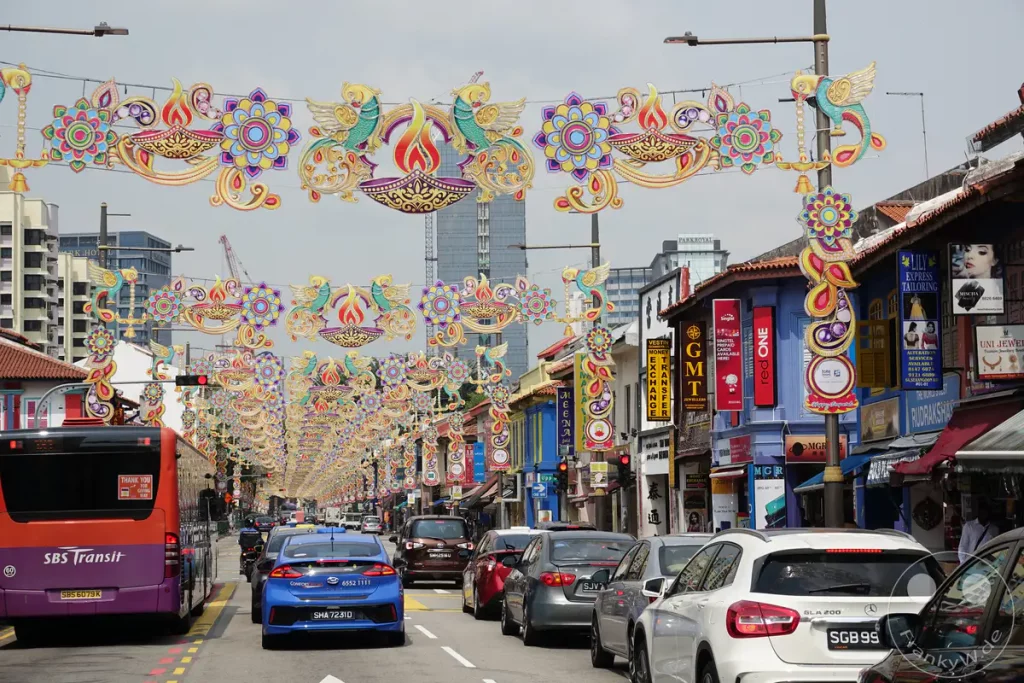 Festliche Lichterketten schmücken die Straße im Viertel Little India in Singapur während der Diwali-Feierlichkeiten. Mehrere Autos und ein SBS Transit Bus sind auf der belebten Straße unterwegs. Die Dekorationen schaffen eine lebendige und festliche Atmosphäre inmitten des städtischen Treibens.