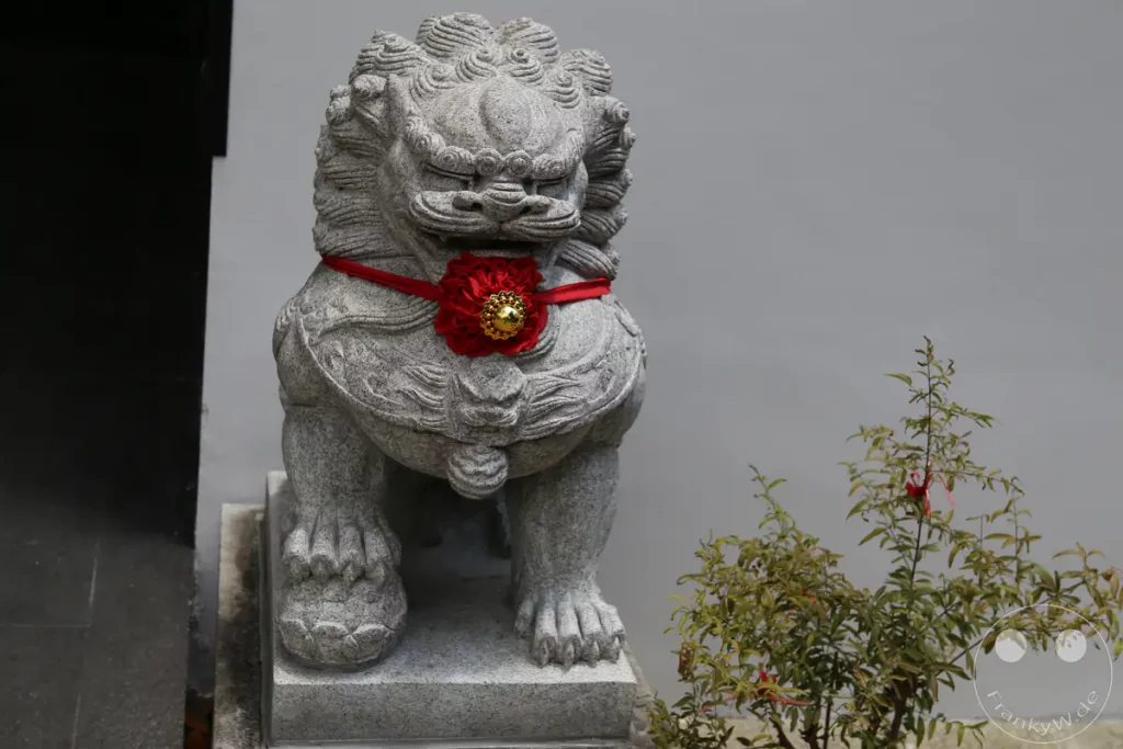 Eine steinerne Löwenstatue, oft als "Fu Dog" bezeichnet, wird mit einem roten Band und einer roten Blume mit goldener Mitte geschmückt. Die Statue steht vor einer schlichten grauen Wand, daneben wächst eine kleine Pflanze mit grünen Blättern und roten Verzierungen. Das Bild strahlt eine Atmosphäre von Schutz und kultureller Tradition aus, die typisch für viele asiatische Kulturen ist.