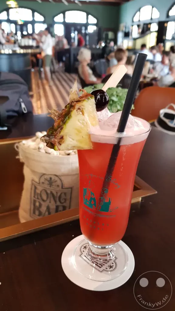 Ein erfrischender Singapore Sling wird im Long Bar serviert und mit einem Stück Ananas und einer Kirsche garniert. Die rote Farbe des Cocktails wirkt ansprechend und lädt zum Genuss ein. Hinter dem Drink sind unscharfe Silhouetten von Gästen zu erkennen, die die Atmosphäre eines belebten Ortes unterstreichen.