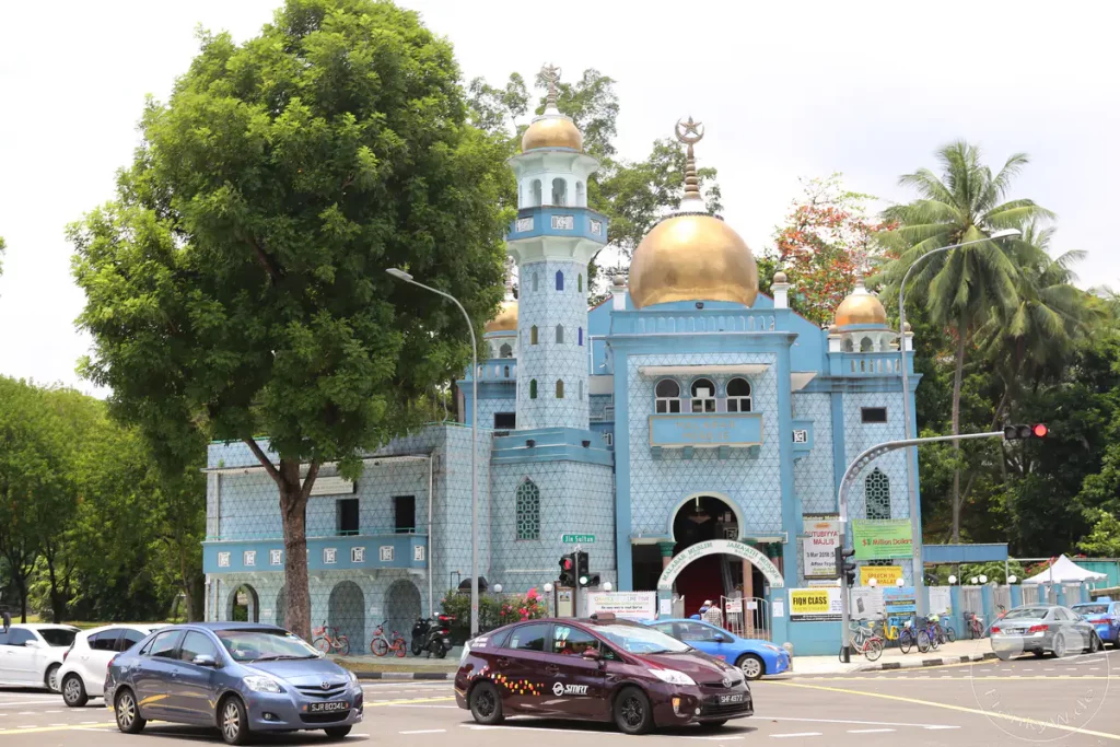 Die Malabar-Moschee in Singapur ist ein auffälliges Gebäude mit türkisfarbener Fassade und goldenen Kuppeln. Vor der Moschee herrscht belebtes Stadtleben mit Autos und Fahrrädern, die die Straße kreuzen. Die Szene strahlt eine friedliche Atmosphäre inmitten der städtischen Geschäftigkeit aus.