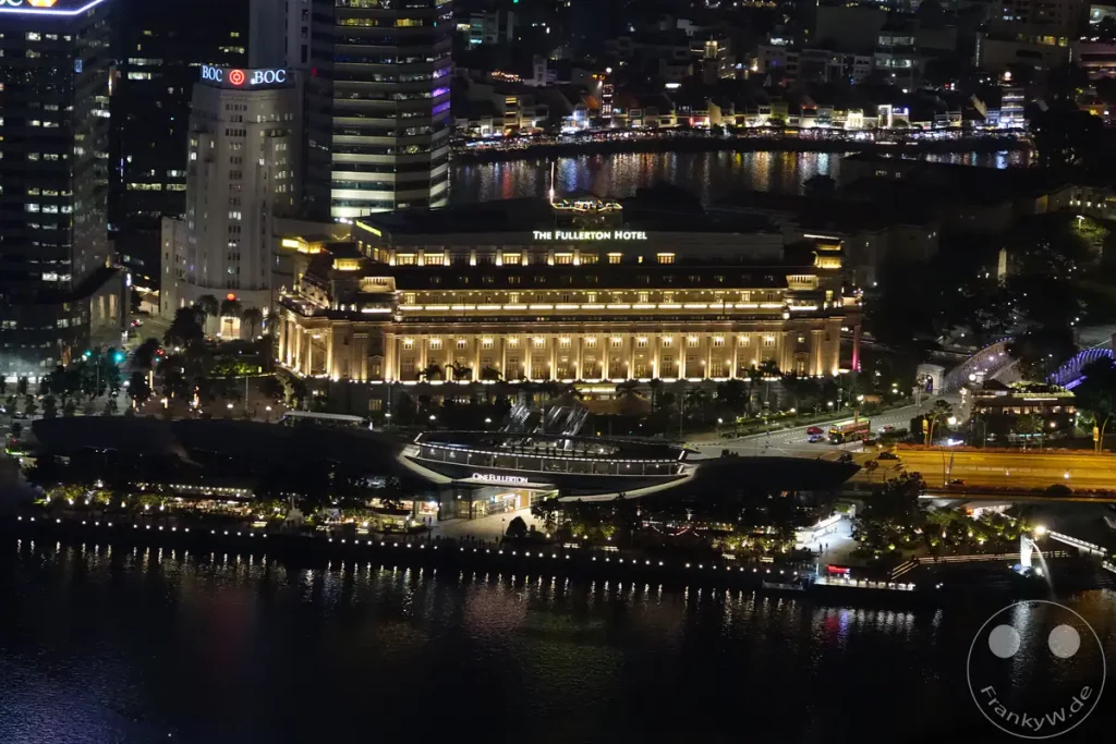 Die Aufnahme zeigt das beleuchtete Fullerton Hotel und die umliegende Stadtlandschaft von Singapur bei Nacht. Die Lichter der Gebäude spiegeln sich im dunklen Wasser des Flusses, was eine stimmungsvolle und glanzvolle Atmosphäre schafft. Eine Fähre liegt am Ufer, während im Hintergrund weitere städtische Strukturen erkennbar sind.