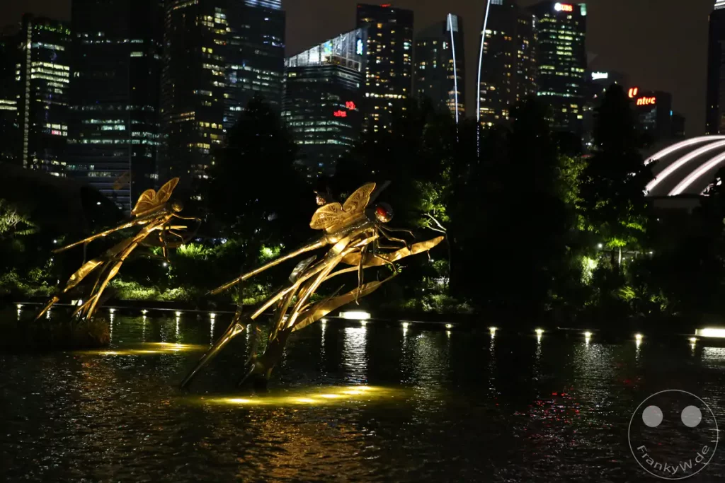 Eine beeindruckende Libellen-Skulptur aus Metall, beleuchtet durch Scheinwerfer, spiegelt sich in einem dunklen Wasserbecken wider. Im Hintergrund erheben sich die beleuchteten Wolkenkratzer der Skyline von Singapur bei Nacht. Die Szene strahlt eine moderne und gleichzeitig mystische Atmosphäre aus.
