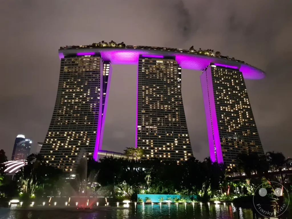 Die beeindruckende Architektur des Marina Bay Sands Hotels in Singapur ist nachts in kräftiges Violett getaucht. Die beleuchteten Fenster der drei Türme spiegeln sich in der ruhigen Wasseroberfläche, während Wolken den Himmel bedecken. Die Szenerie strahlt eine moderne und luxuriöse Atmosphäre aus, die durch die künstliche Beleuchtung hervorgehoben wird.