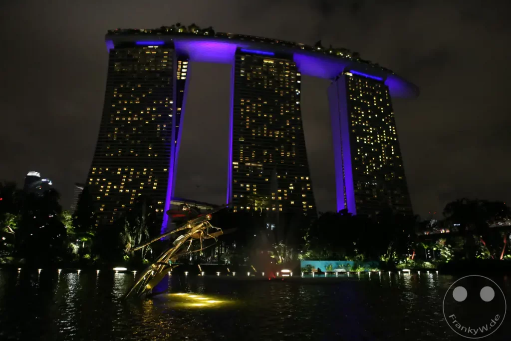 Die ikonische Marina Bay Sands Skyline von Singapur ist nachts beleuchtet und reflektiert im Wasser. Die drei markanten Wolkenkratzer mit der Dachterrasse, die in lila Licht getaucht ist, bilden das Zentrum der Aufnahme. Eine Skulptur im Vordergrund und die Lichter der Stadt erzeugen eine lebendige und moderne Atmosphäre.