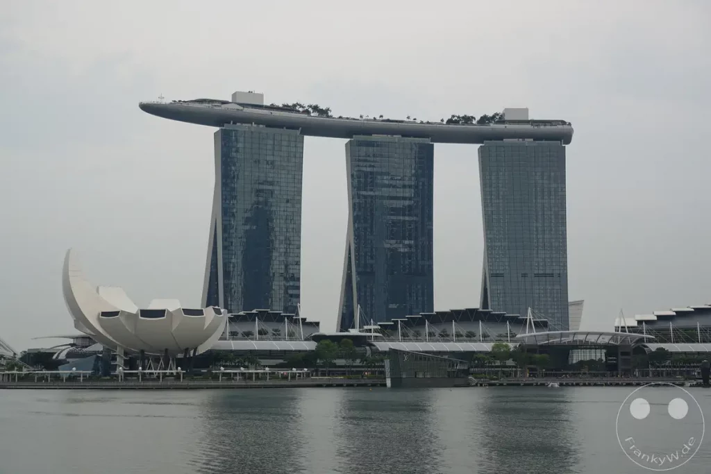 Die ikonische Architektur des Marina Bay Sands Hotels mit seinen drei markanten Türmen, die von einem Dachgarten verbunden sind, dominiert die Skyline von Singapur. Vor dem Hotelkomplex befindet sich eine blütenförmige Struktur, die den Wasserfrontbereich mit modernen Gebäuden ergänzt. Das Bild vermittelt einen Eindruck von der futuristischen und beeindruckenden Stadtlandschaft Singapurs bei trübem Wetter.