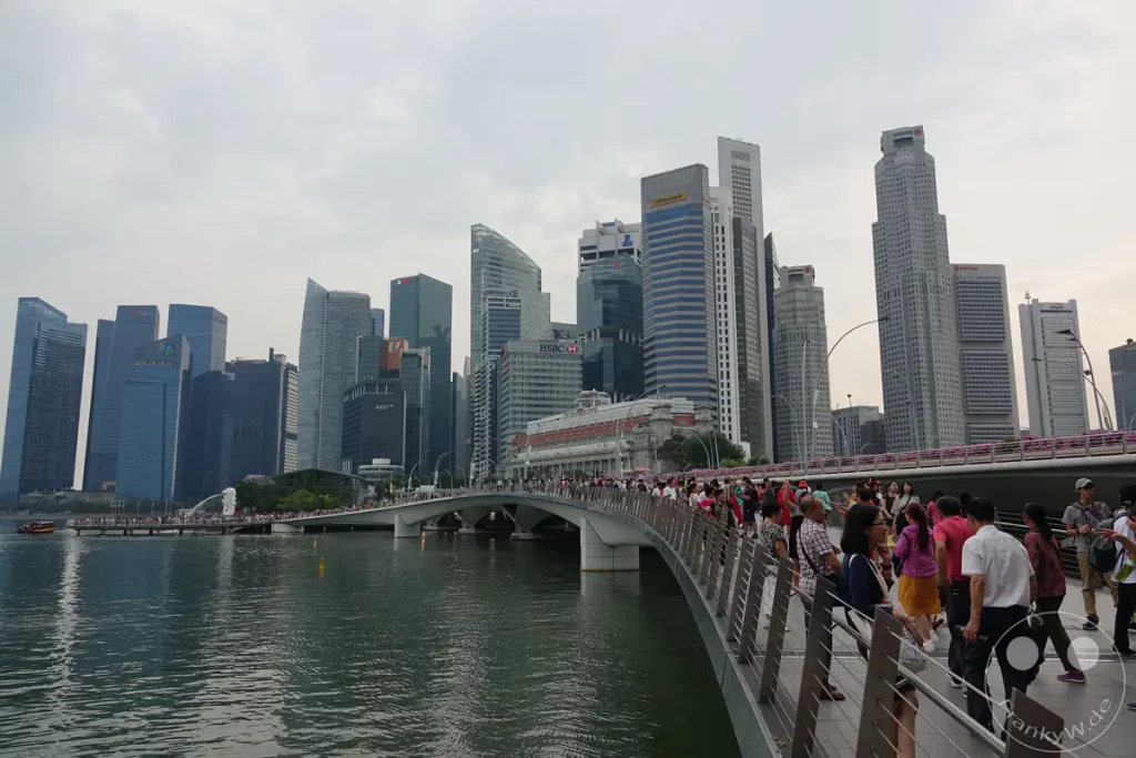 Die Skyline von Singapur erstreckt sich über den Marina Bay, während Menschen über eine geschwungene Fußgängerbrücke spazieren. Die moderne Architektur der Wolkenkratzer bildet einen beeindruckenden Kontrast zum ruhigen Wasser darunter. Die Szene vermittelt eine lebendige und zugleich friedliche Atmosphäre der Stadt.