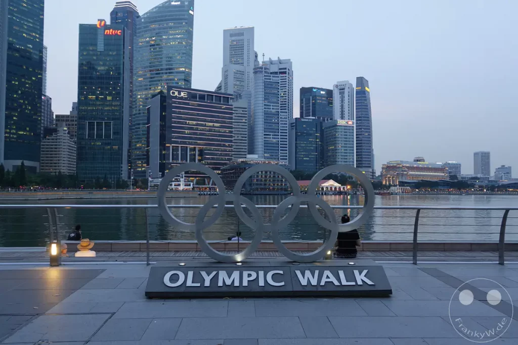 Die Skyline von Singapur spiegelt sich im Wasser, während die ikonischen Olympischen Ringe und das OUE-Gebäude im Vordergrund zu sehen sind. Die beleuchteten Gebäude und die ruhige Atmosphäre der Marina Bay erzeugen eine moderne und dennoch friedliche Szenerie. Zwei Personen sitzen auf der Promenade und genießen den Ausblick auf die Stadt bei Dämmerung.