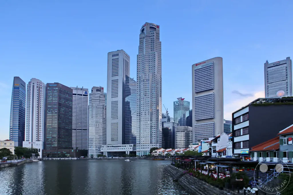 Die beeindruckende Skyline von Singapur spiegelt sich im Wasser des Marina Bay. Zahlreiche Wolkenkratzer säumen das Ufer und bilden ein modernes Stadtbild. Entlang des Flusses laden Restaurants zum Verweilen ein und erzeugen eine lebendige Atmosphäre.