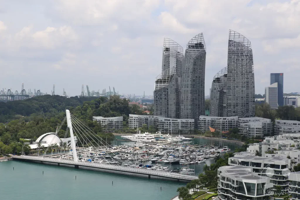 Eine weitläufige Ansicht von Singapurs Marina Bay, mit einem überfüllten Yachthafen im Vordergrund, der unzählige Boote beherbergt. Dahinter ragen markante, moderne Wolkenkratzer in den Himmel, während grüne Hügel und weiter entfernte Containerkräne die urbane Landschaft ergänzen. Der Himmel ist mit zarten Wolken bedeckt, was der Szene eine ruhige und doch dynamische Atmosphäre verleiht.