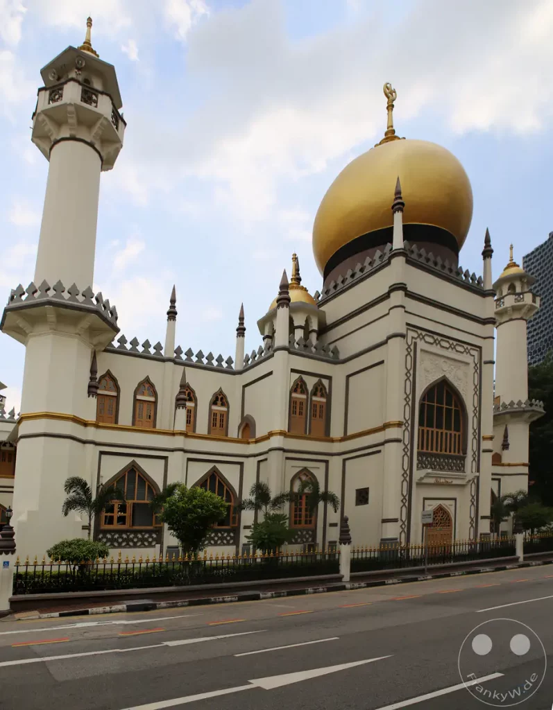 Die Masjid Sultan in Singapur besticht durch ihre beeindruckende Architektur mit der markanten goldenen Kuppel und dem schlanken Minarett. Das Gebäude spiegelt eine Mischung aus traditionellen und modernen islamischen Baustilen wider. Die friedliche Atmosphäre wird durch die umliegende Vegetation und den blauen Himmel unterstrichen.
