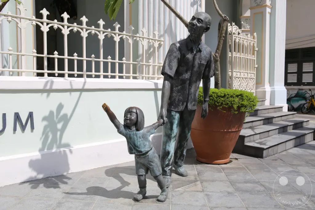 Eine Bronzeskulptur vor einem Gebäude in Singapur stellt einen älteren Mann dar, der ein Kind an der Hand führt. Das Denkmal symbolisiert die Weitergabe von Wissen und Erinnerungen. Die Szene vermittelt eine warme und nostalgische Atmosphäre unter dem Schatten von Bäumen.