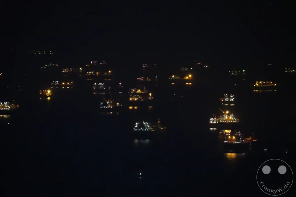 Mehrere beleuchtete Schiffe und Boote liegen im Hafen von Singapur in der Nacht vor Anker. Die Lichter der Schiffe spiegeln sich im dunklen Wasser und erzeugen eine stimmungsvolle Atmosphäre. Eine belebte Wasserstraße bei Dunkelheit ist zu erkennen.