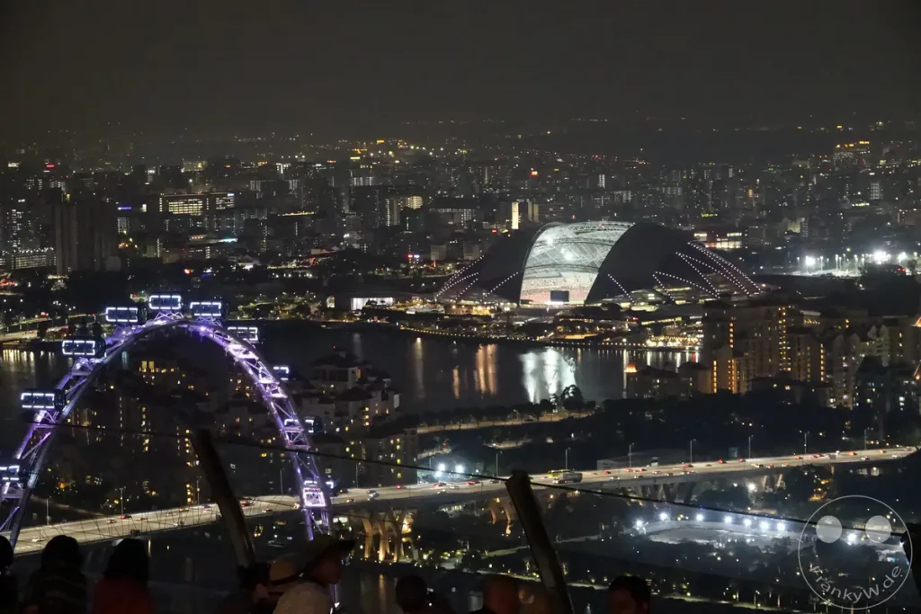 Die nächtliche Skyline von Singapur erstreckt sich unter einem dämmrigen Himmel. Beleuchtete Wahrzeichen wie der Singapore Flyer und die beeindruckende Architektur des Marina Bay Sands Stadions dominieren die Szenerie. Die Lichter der Stadt spiegeln sich im Wasser und schaffen eine lebendige und faszinierende Atmosphäre.