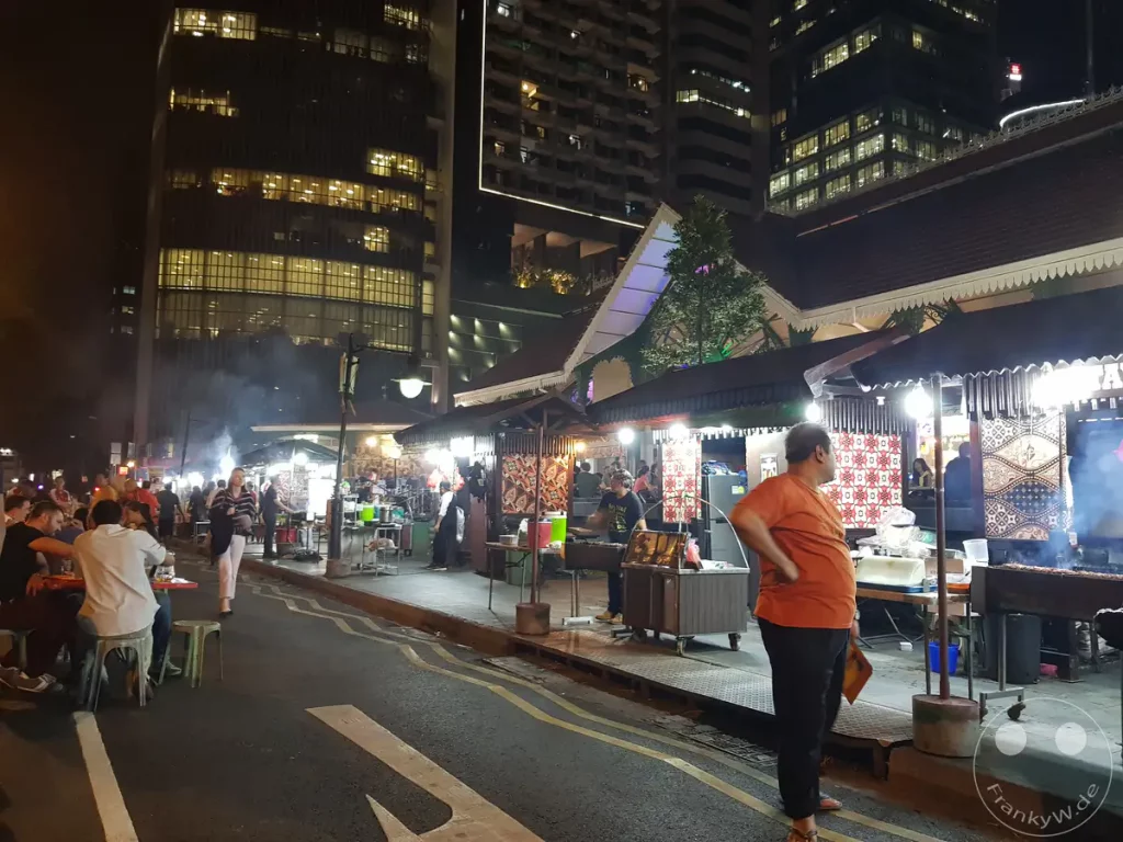 In Singapur herrscht auf einem belebten Nachtmarkt reges Treiben, wo Menschen an Ständen speisen und sich unterhalten. Die beleuchteten Gebäude im Hintergrund bilden einen modernen Kontrast zu den traditionellen Essensständen und der ausgelassenen Atmosphäre. Rauch von den Grillstationen und das gedämpfte Licht schaffen eine lebendige und einladende Stimmung.