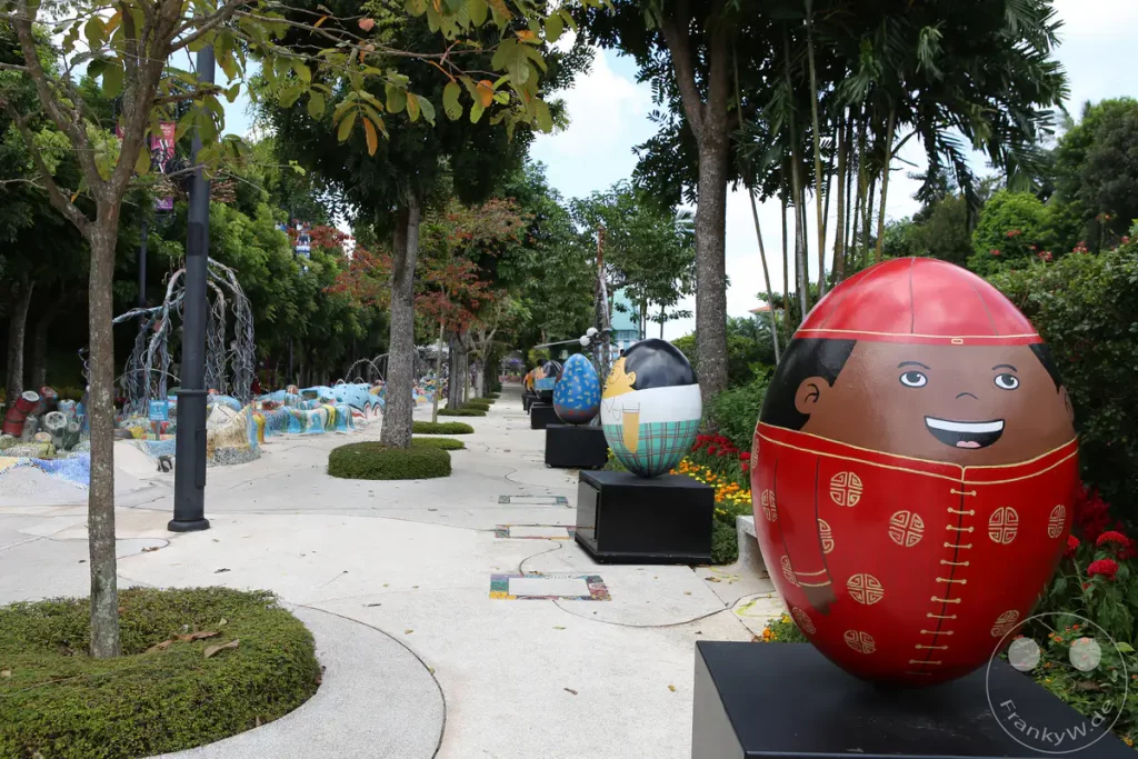 Eine Reihe von farbenfrohen, eiförmigen Skulpturen, die menschliche Gesichter darstellen, säumen einen Weg in einem Park in Singapur. Die Skulpturen sind mit traditionellen Mustern verziert und stehen auf schwarzen Sockeln. Im Hintergrund sind üppige Bäume und Sträucher zu sehen, was eine fröhliche und festliche Atmosphäre schafft.