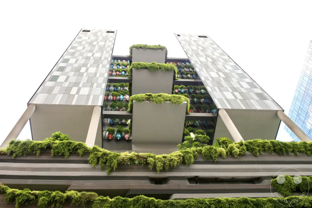 Eine ungewöhnliche und moderne Architektur, die vertikale Gärten in ihre Fassade integriert, dominiert den Blickpunkt. Die üppige grüne Vegetation kontrastiert stark mit den geometrischen Mustern der Gebäudestruktur und des angrenzenden Wolkenkratzers. Das Bild vermittelt ein Gefühl von urbaner Nachhaltigkeit und futuristischem Design in Singapur.