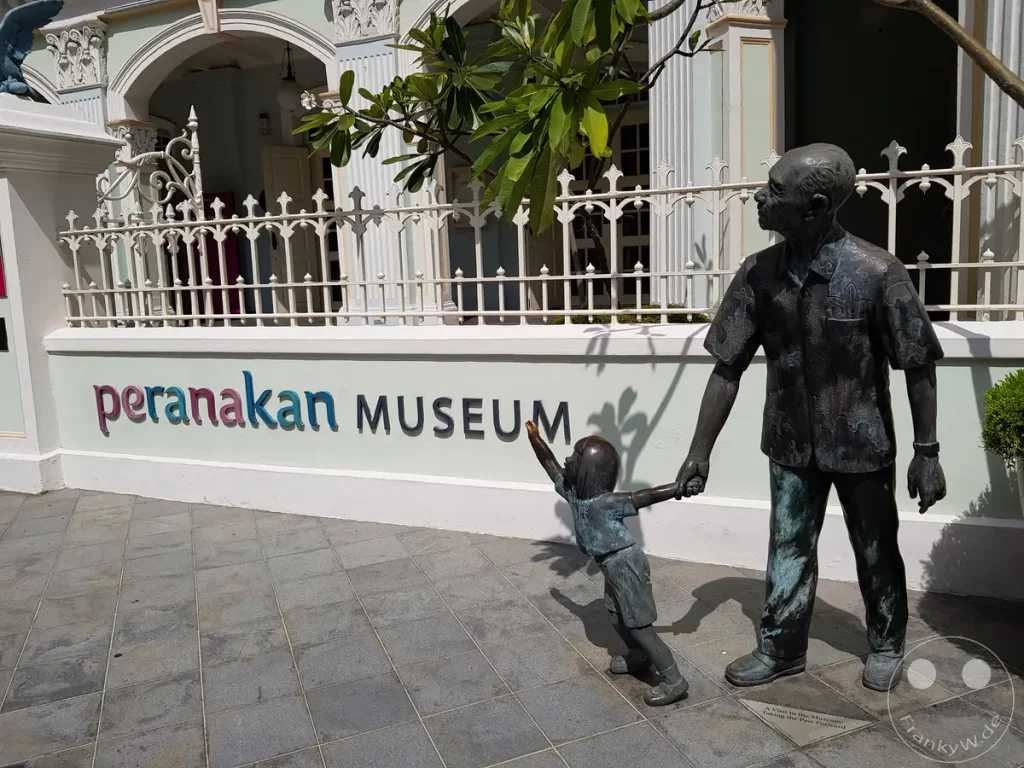 Eine Bronzeskulptur eines Mannes und eines Kindes steht vor dem Peranakan Museum in Singapur. Die Skulptur mit dem Titel "A Visit to the Museum: Making the Past Forward" (Ein Besuch im Museum: Die Vergangenheit vorwärts gestalten) symbolisiert die Verbindung von Generationen und die Weitergabe von Traditionen. Die lebhafte Darstellung vermittelt ein Gefühl von Wärme und kulturellem Erbe.