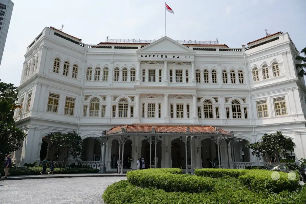 Das weltberühmte Raffles Hotel in Singapur präsentiert sich in seiner eleganten kolonialen Architektur unter einem hellen Himmel. Vor dem Gebäude lädt eine gepflegte Grünanlage mit Sträuchern und Bäumen zum Verweilen ein. Menschen bewegen sich auf dem Gehweg vor der beeindruckenden Fassade, während Autos diskret am Rand parken und zur Szenerie beitragen.
