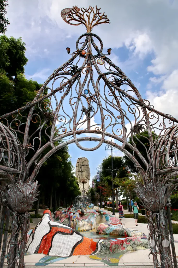 Ein kunstvoller, metallener Torbogen mit floralen Elementen rahmt den Blick auf einen farbenfrohen, mosaikverzierten Brunnen in Singapur. Im Hintergrund ist die ikonische Merlion-Statue zu sehen, umgeben von üppigem Grün und blauen Himmel mit Wolken. Die Szene vermittelt eine lebendige und märchenhafte Atmosphäre in einem tropischen Urlaubsort.