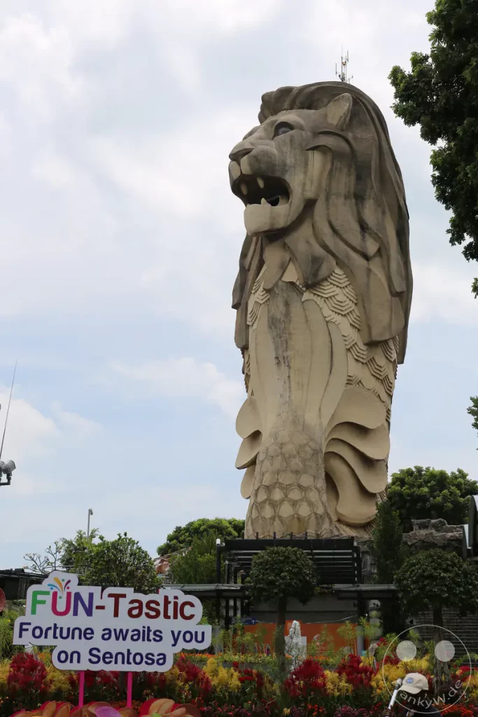 Die imposante Merlion-Statue in Singapur, ein Symbol der Stadt, ragt majestätisch in den Himmel. Bunte Blumenarrangements und üppige Vegetation umrahmen das Wahrzeichen und schaffen eine lebendige und einladende Atmosphäre. Das Foto fängt die touristische Anziehungskraft von Sentosa Island ein und verspricht ein aufregendes Erlebnis für Besucher.