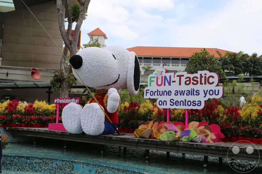 Eine riesige Snoopy-Figur aus Blumen sitzt auf einer Holzplattform vor einem Gebäude mit orangeroten Ziegeldach. Vor der Figur steht ein Schild mit der Aufschrift "FUN-Tastic" und "Fortune awaits you on Sentosa". Die Szenerie ist mit bunten Blumen geschmückt und vermittelt eine festliche und einladende Atmosphäre, die zu einem Besuch auf Sentosa einlädt.