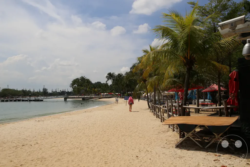 Ein sonniger Tag am Palawan Beach auf Sentosa, Singapur. Heller Sandstrand lädt zum Spazierengehen ein, während im Hintergrund Palmen Schatten spenden und das ruhige Meer zum Baden einlädt. Eine entspannte tropische Atmosphäre herrscht an diesem beliebten Ausflugsziel.