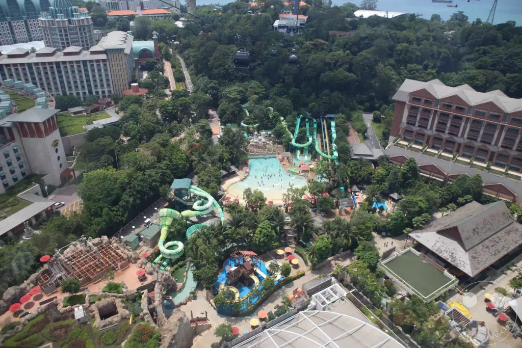 Luftaufnahme eines tropischen Wasserparks mit zahlreichen Wasserrutschen und Pools, umgeben von üppiger Vegetation. Menschen genießen das Wasservergnügen in einem wellenförmigen Pool, während im Hintergrund Hotels und eine Seilbahn zu sehen sind. Die Szene strahlt Urlaubsfreude und Erholung in einer sonnigen Umgebung aus.
