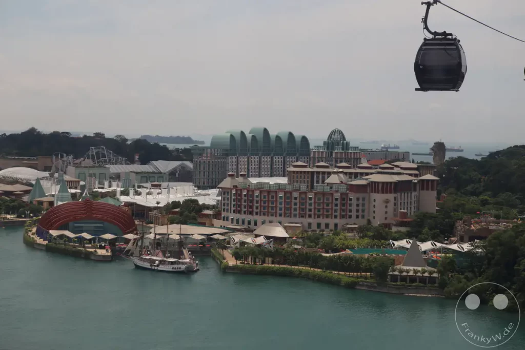 Eine weite Ansicht von Sentosa Island in Singapur mit einem Blick auf eine Seilbahnkabine im Vordergrund. Ein großes Resort mit interessanter Architektur liegt am Wasser, während Schiffe auf dem Meer im Hintergrund sichten sind. Die Szene vermittelt ein Gefühl von Urlaub und entspannter Atmosphäre an einem tropischen Ort.