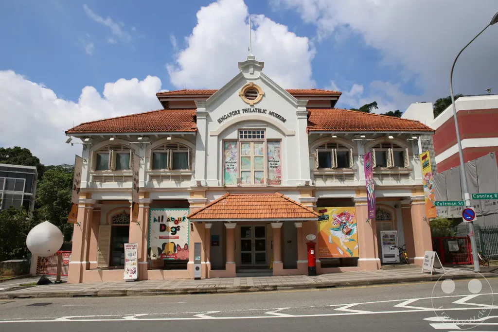 Das Singapore Philatelic Museum präsentiert sich in einem historischen Gebäude mit klassischer Architektur. Vor dem Museum werben farbenfrohe Banner für aktuelle Ausstellungen. Die Szene strahlt eine ruhige, städtische Atmosphäre unter einem bewölkten Himmel aus.