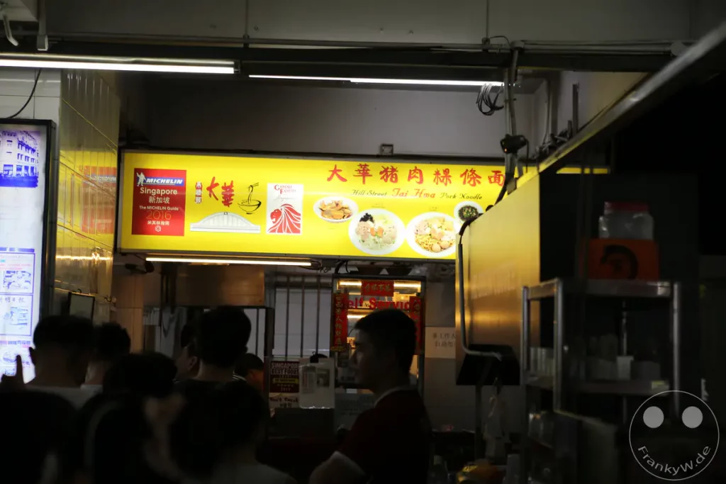 Ein beleuchtetes Schild über einem Imbissstand in Singapur zeigt die Auszeichnung des Michelin Guide 2016 und wirbt für "Hill Street Tai Hwa Pork Noodle". Die Szene wirkt lebhaft, da sich im Vordergrund einige Personen befinden, die auf ihre Bestellung warten oder sich unterhalten. Das helle Leuchten des Schildes kontrastiert mit der Dunkelheit des Hintergrunds und erzeugt eine typische Atmosphäre eines belebten Nachtmarktes oder einer Essensstraße.