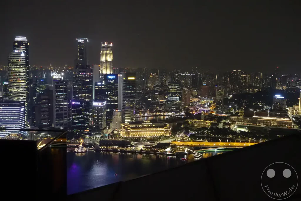 Die beleuchtete Skyline von Singapur erstreckt sich bei Nacht über den Wasserspiegel des Marina Bay. Zahlreiche Wolkenkratzer und historische Gebäude spiegeln sich in der Dunkelheit und erzeugen eine beeindruckende städtische Atmosphäre. Kleine Boote kreuzen auf dem Wasser und tragen zur lebendigen Szenerie bei.