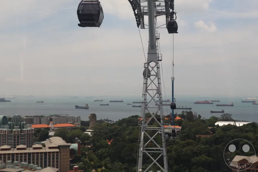 Eine Seilbahn überquert den Horizont von Singapur, mit zahlreichen Frachtschiffen, die vor der Küste ankern. Die Stadtlandschaft mit ihren modernen Gebäuden und üppigen grünen Hügeln ist im Hintergrund zu sehen. Das Bild vermittelt ein Gefühl von urbaner Geschäftigkeit und der Bedeutung Singapurs als wichtiger Seehafen.