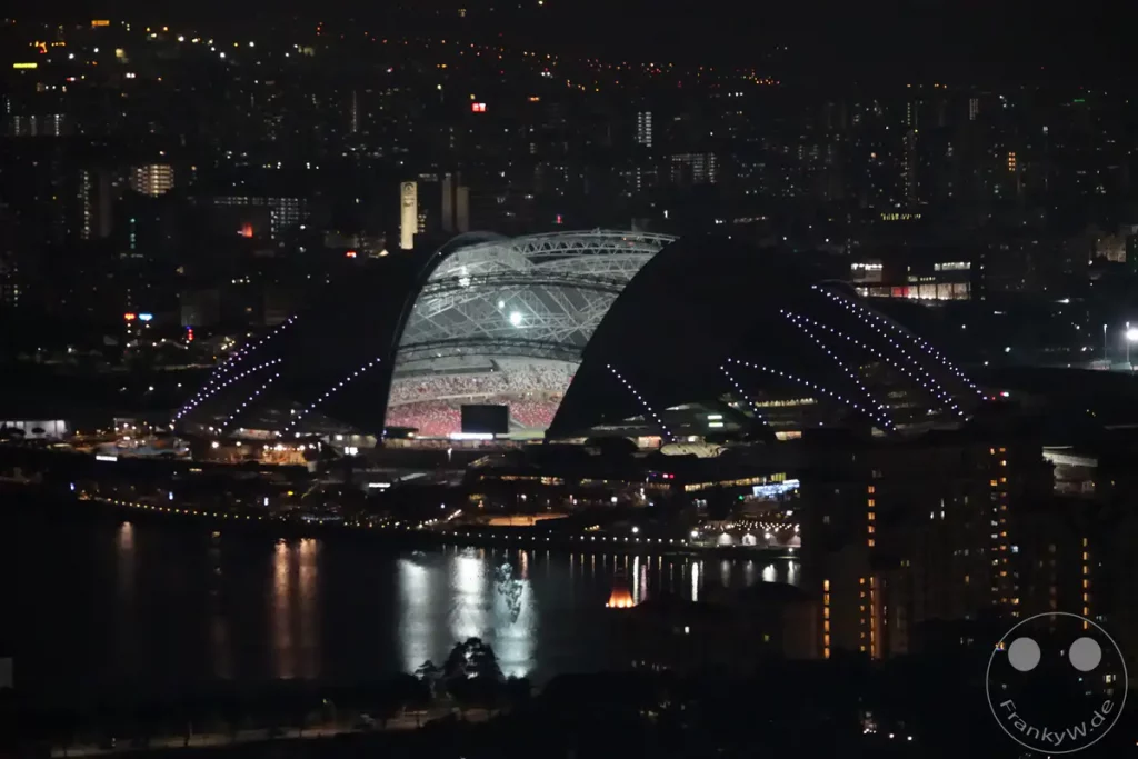 Eine nächtliche Stadtlandschaft von Singapur, dominiert von der imposanten Architektur eines Stadions mit seinem auffälligen Dach und den beleuchteten Sitzreihen. Das umliegende Stadtgebiet zeigt ein glitzerndes Lichtermeer, das sich auf dem dunklen Wasser des Hafens spiegelt. Die Szene vermittelt eine lebendige und dynamische Atmosphäre der Metropole bei Nacht.