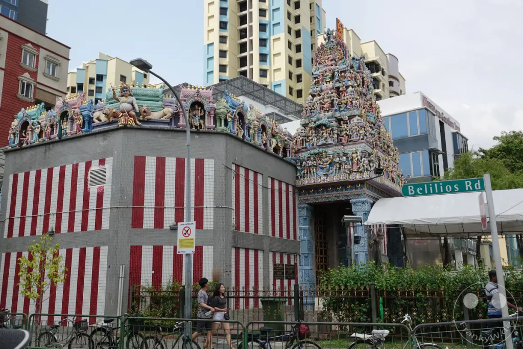 Die farbenprächtige Architektur eineshinduistischen Tempels in Singapur wird durch seine detailreichen Skulpturen und Figuren hervorgehoben. Moderne Hochhäuser bilden einen deutlichen Kontrast zur traditionellen Bauweise des Gotteshauses. Menschen gehen an einer belebten Straße vorbei, während Fahrräder am Zaun abgestellt sind.