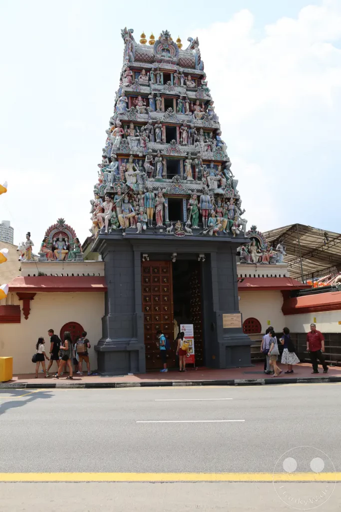 Der Sri Veeramakaliamman Tempel in Singapur präsentiert sich mit seiner detailreichen, farbenfrohen Architektur, die von zahlreichen Skulpturen und Verzierungen geschmückt ist. Eine Gruppe von Menschen bewegt sich am Eingang des Tempels, was auf seine Funktion als lebendiger Ort der Anbetung und des kulturellen Austauschs hinweist. Das Bild vermittelt eine Atmosphäre der Geschäftigkeit und spirituellen Bedeutung im Herzen von Singapur.