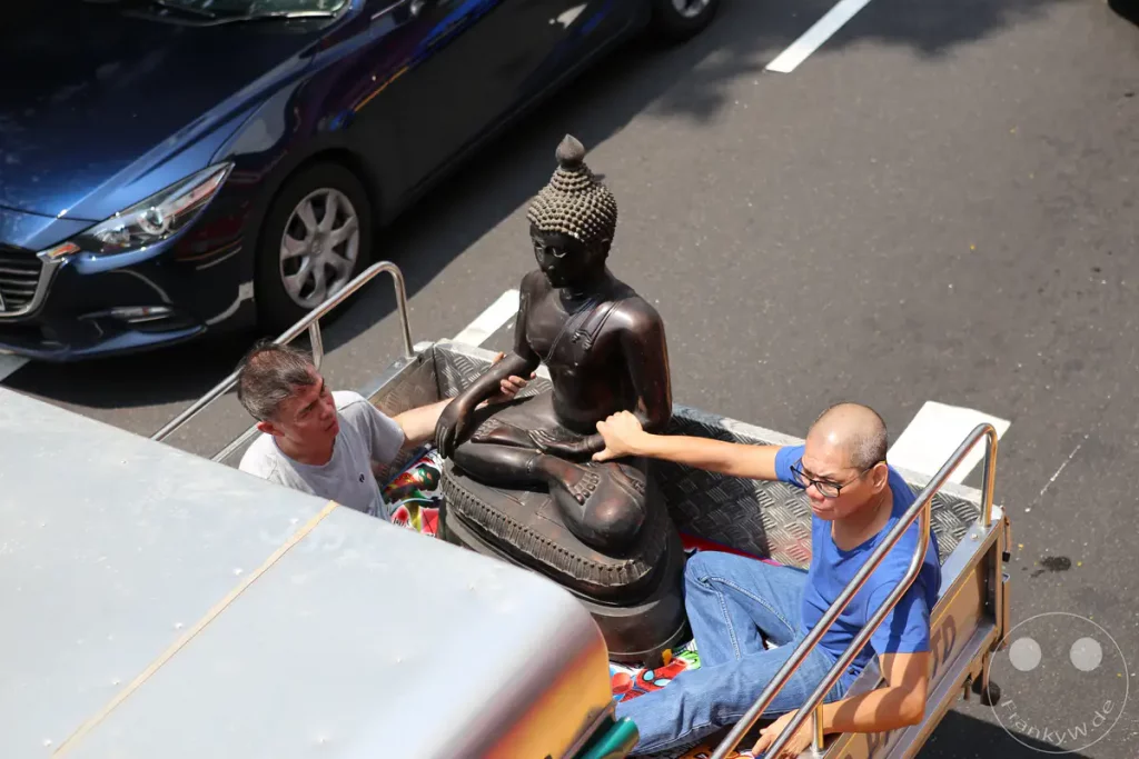 Zwei Männer transportieren eine sitzende Buddha-Statue in einem offenen Fahrzeug. Sie fahren auf einer Straße, wobei im Hintergrund ein Auto zu sehen ist. Die Szene wirkt friedlich, trotz des städtischen Umfelds.