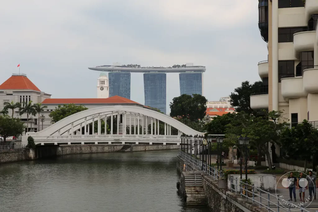 Das Bild zeigt eine Szene am Flussufer in Singapur mit einer markanten weißen Brücke im Vordergrund. Im Hintergrund sind die ikonischen Türme des Marina Bay Sands Hotels und ein Uhrenturm zu sehen. Die Atmosphäre des Bildes ist ruhig und urban, typisch für die moderne Skyline der Stadt.