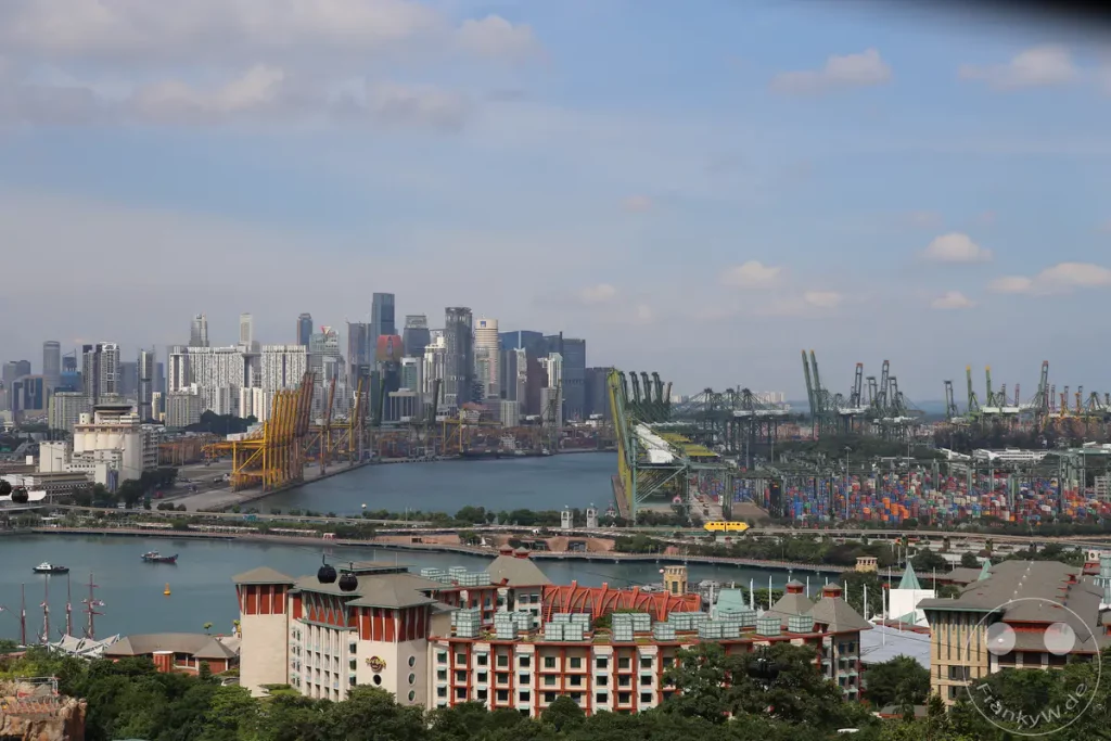 Ein weitläufiger Blick über die Skyline von Singapur, die eine Mischung aus modernen Wolkenkratzern und geschäftigen Hafenanlagen zeigt. Große Containerkräne und gestapelte Frachtcontainer dominieren den unteren Teil des Bildes und zeugen von der industriellen Aktivität der Stadt. Im Vordergrund sind einige Gebäude und üppige grüne Bäume zu sehen, während im Himmel vereinzelte Wolken für eine leichte Stimmung sorgen.