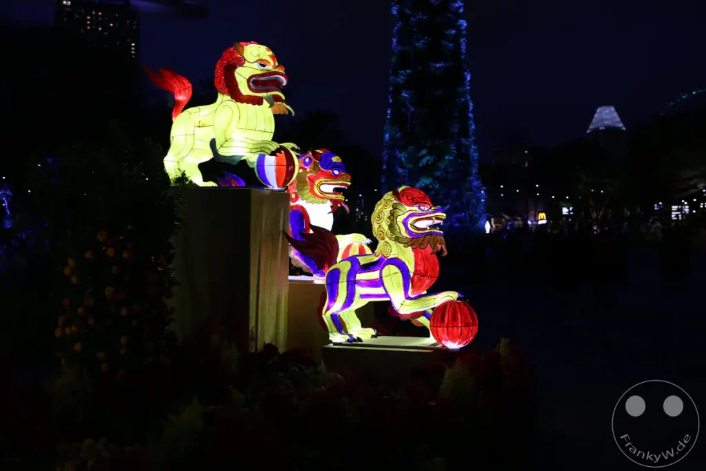 Bunte, leuchtende Laternen stellen traditionelle chinesische Löwenfiguren dar, die bei Nacht beleuchtet werden. Die aufwendigen Kunstwerke stehen inmitten von dunklen Bäumen und Büschen, die mit kleinen Lichtern geschmückt sind. Im Hintergrund sind die Lichter von Stadtgebäuden erkennbar, was eine festliche Atmosphäre erzeugt.