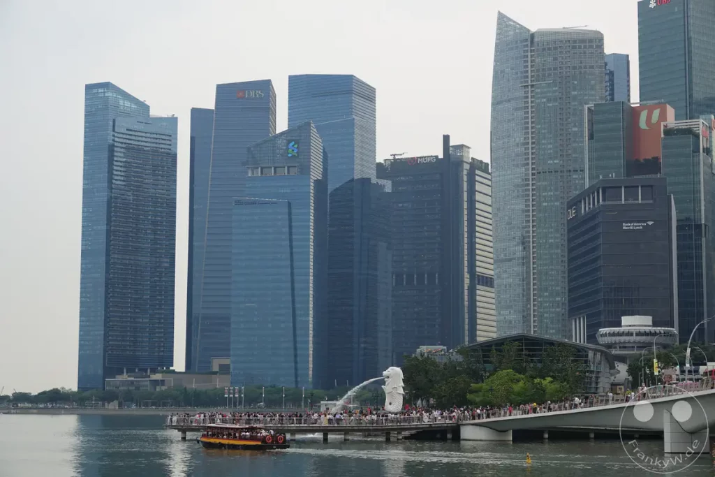 Die beeindruckende Skyline von Singapur wird von modernen Wolkenkratzern dominiert, die sich im Wasser spiegeln. In der Vordergrundszene zieht ein Boot durch den Fluss, während Menschen auf einer Brücke entlanggehen und die Aussicht genießen. Die Atmosphäre ist lebhaft und zeugt von der dynamischen Metropole.
