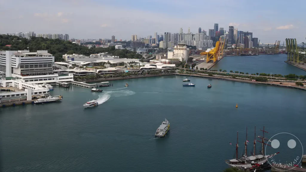 Ein Panoramablick auf den geschäftigen Hafen von Singapur, der die moderne Stadtarchitektur mit dem lebhaften Schiffsverkehr im Wasser verbindet. Mehrere Boote, Fähren und ein Segelschiff sind auf dem türkisfarbenen Wasser unterwegs, während sich im Hintergrund eine beeindruckende Skyline abzeichnet. Die Szene vermittelt ein Gefühl von urbaner Dynamik und maritimer Aktivität an einem sonnigen Tag.