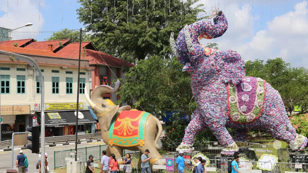 Die Aufnahmen zeigen zwei aufwendig gestaltete Elefanten-Skulpturen im Freien in Singapur. Eine der Skulpturen ist mit bunten Blumen bedeckt, während die andere ein traditionelleres, bemaltes Design aufweist. Die Szenerie strahlt eine festliche Atmosphäre aus, die wahrscheinlich mit einem kulturellen Ereignis oder einem Festival verbunden ist.