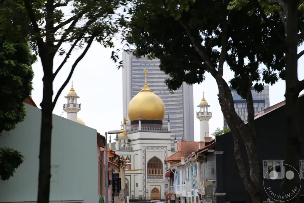 Die Sultan-Moschee mit ihrer markanten goldenen Kuppel und den schlanken Minaretten ist ein Wahrzeichen Singapurs. Sie ist umgeben von der historischen Architektur der Arab Street, einer pulsierenden Gegend mit Geschäften und Boutiquen. Die Szene vermittelt einen Eindruck von kultureller Vielfalt und historischem Erbe inmitten des modernen Stadtbildes von Singapur.