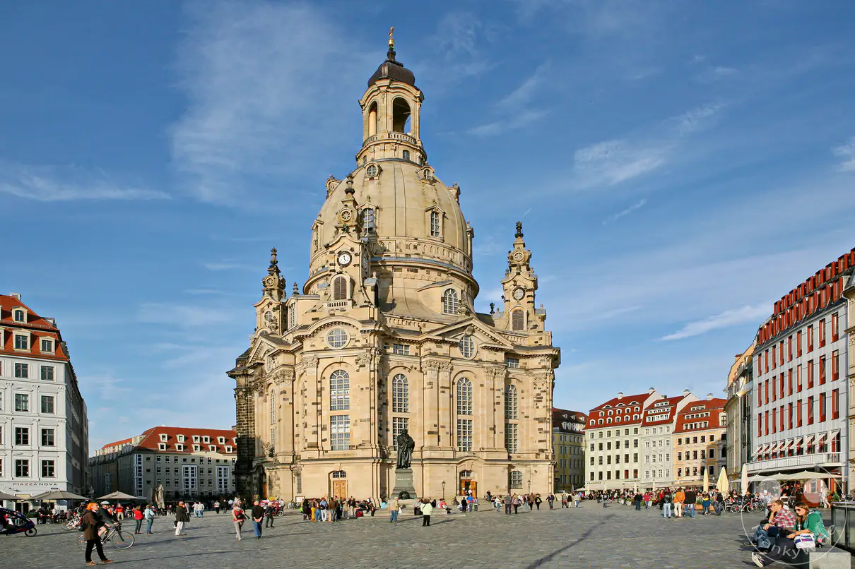 Die imposante Frauenkirche dominiert den weiten, gepflasterten Platz in Dresden, umgeben von historischen Gebäuden unter einem strahlend blauen Himmel. Menschen flanieren und verweilen auf dem Platz, was eine lebendige und friedliche Atmosphäre schafft. Die barocke Architektur und die sonnige Szenerie verleihen dem Ort eine beeindruckende historische Kulisse.