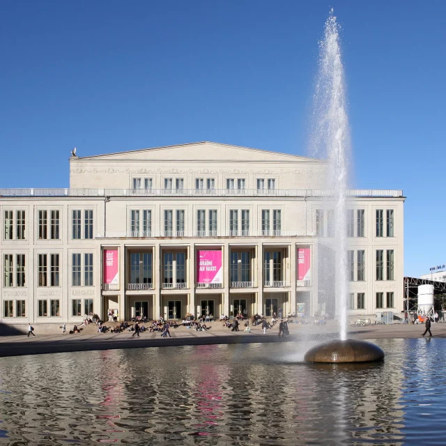 Das Leipziger Opernhaus bildet den beeindruckenden Hintergrund für einen belebten Platz mit einem großen Brunnen im Vordergrund. Menschen sitzen und spazieren auf den Treppen und im Bereich um das Wasser und genießen den sonnigen Tag in Leipzig, Deutschland. Die klassische Architektur des Opernhauses steht im Kontrast zum modernen Brunnen und dem klaren blauen Himmel.
