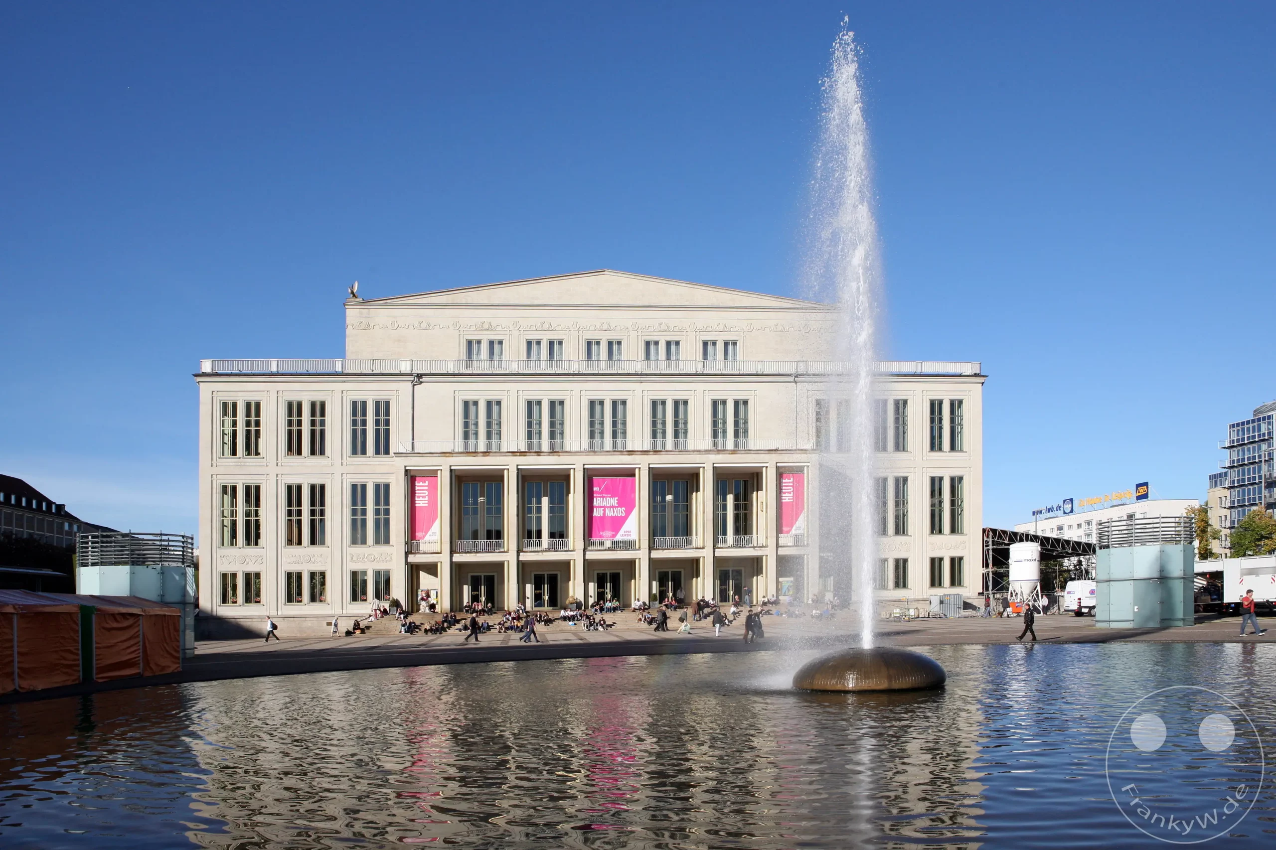 Das Leipziger Opernhaus bildet den beeindruckenden Hintergrund für einen belebten Platz mit einem großen Brunnen im Vordergrund. Menschen sitzen und spazieren auf den Treppen und im Bereich um das Wasser und genießen den sonnigen Tag in Leipzig, Deutschland. Die klassische Architektur des Opernhauses steht im Kontrast zum modernen Brunnen und dem klaren blauen Himmel.