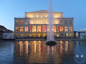 Das Leipziger Opernhaus am Augustusplatz erstrahlt in warmem Abendlicht, während eine Fontäne mitten im Wasserbecken aufsteigt. Die beleuchteten Fenster und Banner spiegeln sich im ruhigen Wasser und schaffen eine festliche und zugleich ruhige Atmosphäre in der deutschen Stadt. Im Vordergrund sind die Lichter der Stadt und des Gebäudes auf der Wasseroberfläche reflektiert, was dem Bild eine zusätzliche Tiefe verleiht.