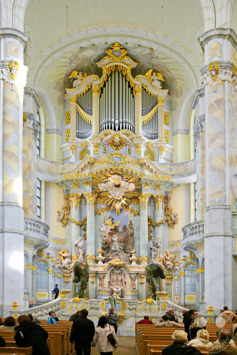 Die prächtige Innenausstattung der Frauenkirche in Dresden ist von barocker Pracht geprägt, mit einer beeindruckenden Orgel und einem reich verzierten Altarraum. Gläubige und Besucher versammeln sich auf den Bänken und nehmen die erhabene Atmosphäre des Gotteshauses wahr. Marmorsäulen, goldene Verzierungen und aufwendige Skulpturen schaffen ein festliches und spirituelles Ambiente.