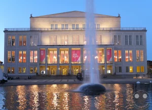 Das Leipziger Opernhaus am Augustusplatz erstrahlt im Abendlicht, während ein Springbrunnen im Vordergrund Wasserfontänen in die Höhe schickt. Die Lichter des Gebäudes spiegeln sich auf der Wasseroberfläche und erzeugen eine atmosphärische Szene in der Stadt. Servicefahrzeuge stehen am Rand des Platzes und deuten auf eine Veranstaltung oder einen Einsatz hin.